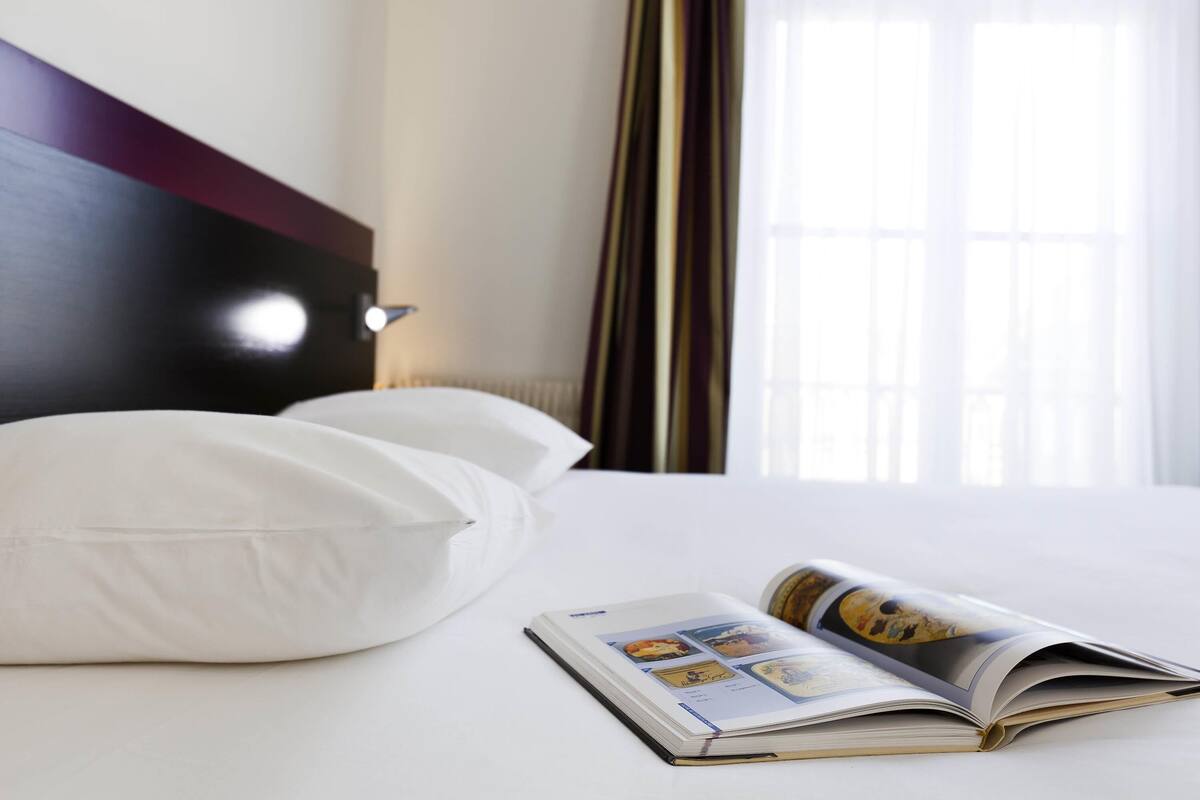 A hotel room with a bed, pillows, a book, and a window with curtains.