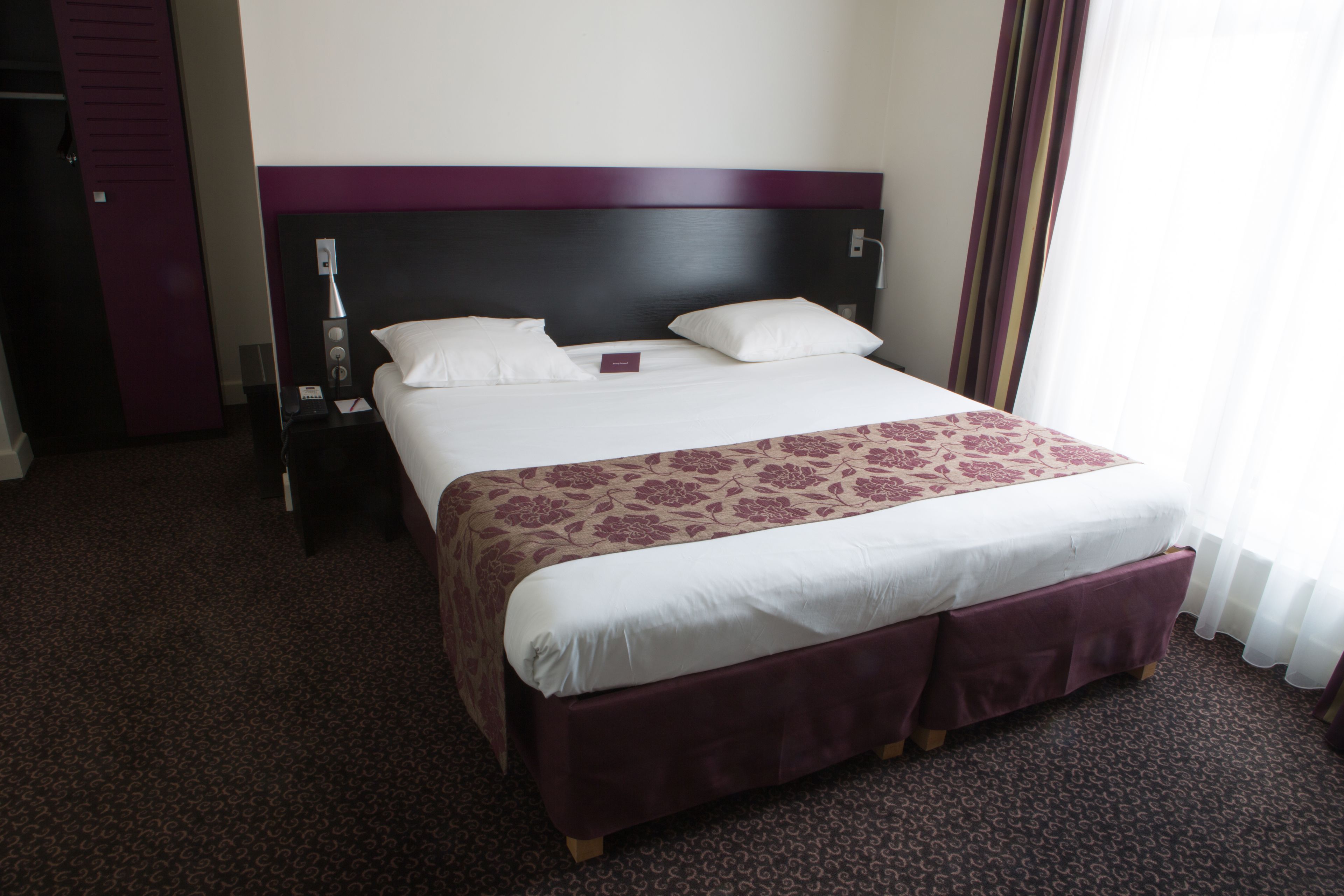 Habitación de hotel con una cama grande, cabecero, mesita de noche y ventana con cortinas.