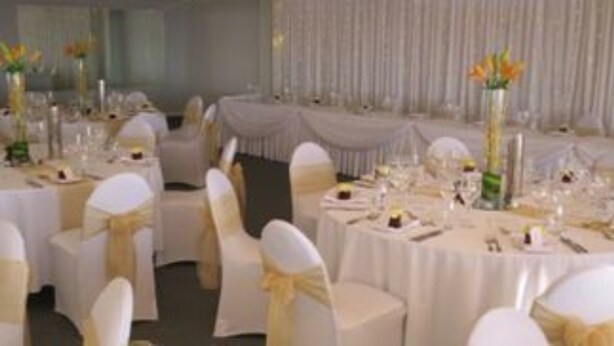 A banquet hall with round tables covered in white tablecloths, each set with white chairs adorned with yellow sashes. - Ocean Beach Hotel (Cottesloe)