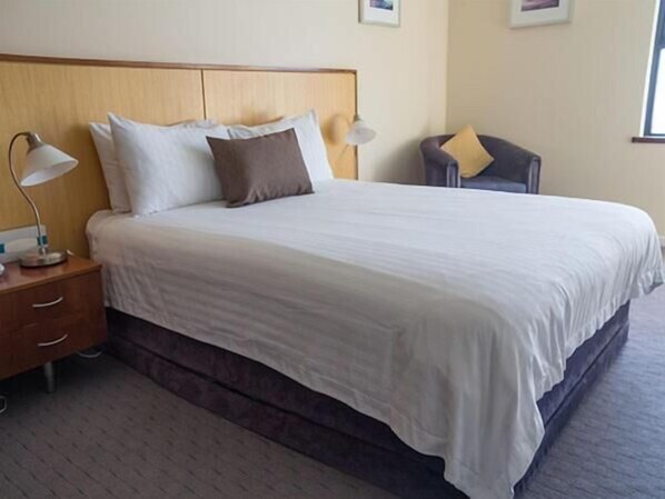 A neatly made bed with white linens and a wooden headboard, a bedside table with a lamp, and a chair with a cushion.