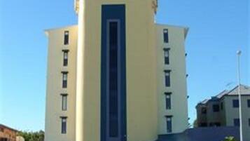 Un edificio de varios pisos con una banda vertical azul en el centro, rodeado de césped y coches aparcados.