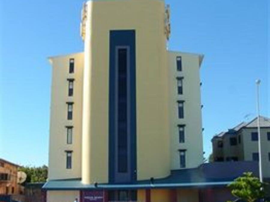Un edificio de varios pisos con una banda vertical azul en el centro, rodeado de césped y coches aparcados.