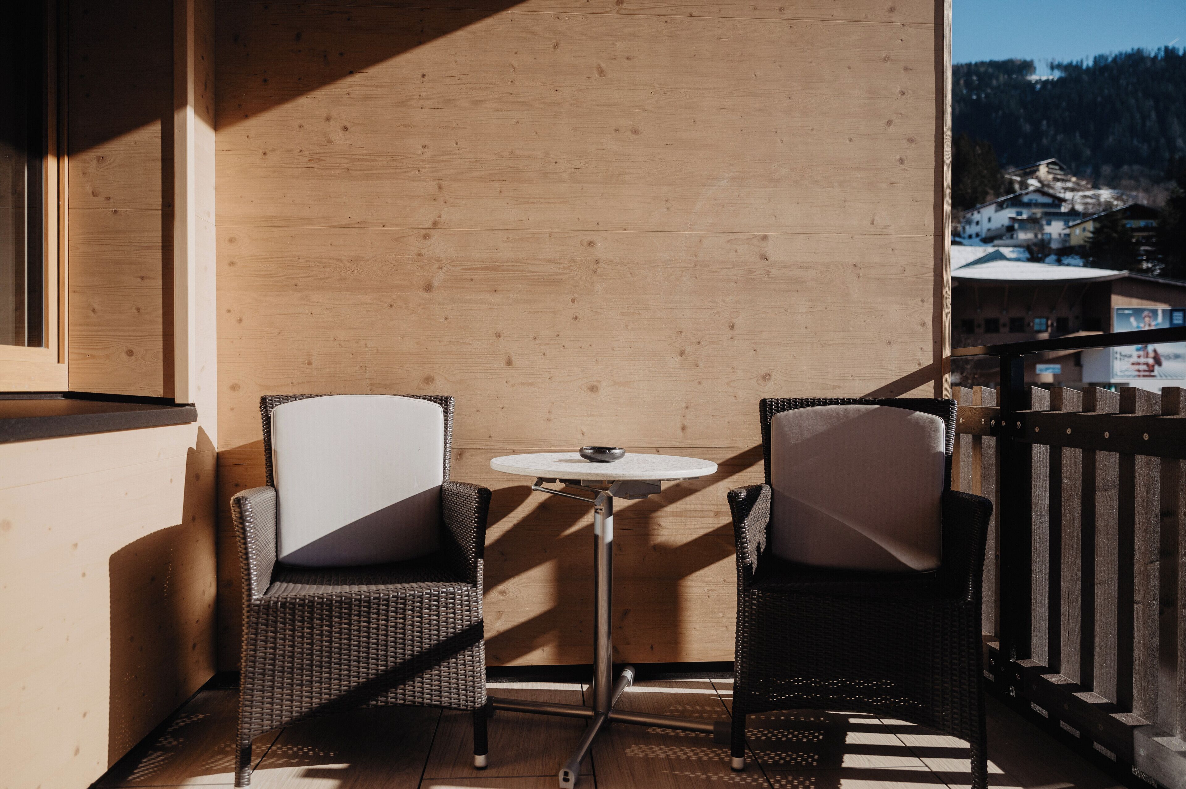 A balcony with wicker chairs and a small round table, overlooking a mountainous village.