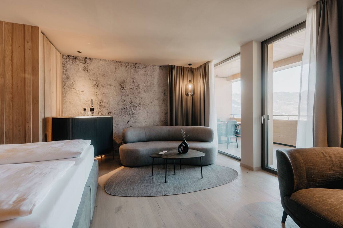 A modern hotel room with a large bed, a sofa, a small round table, and a view of the outdoors.
