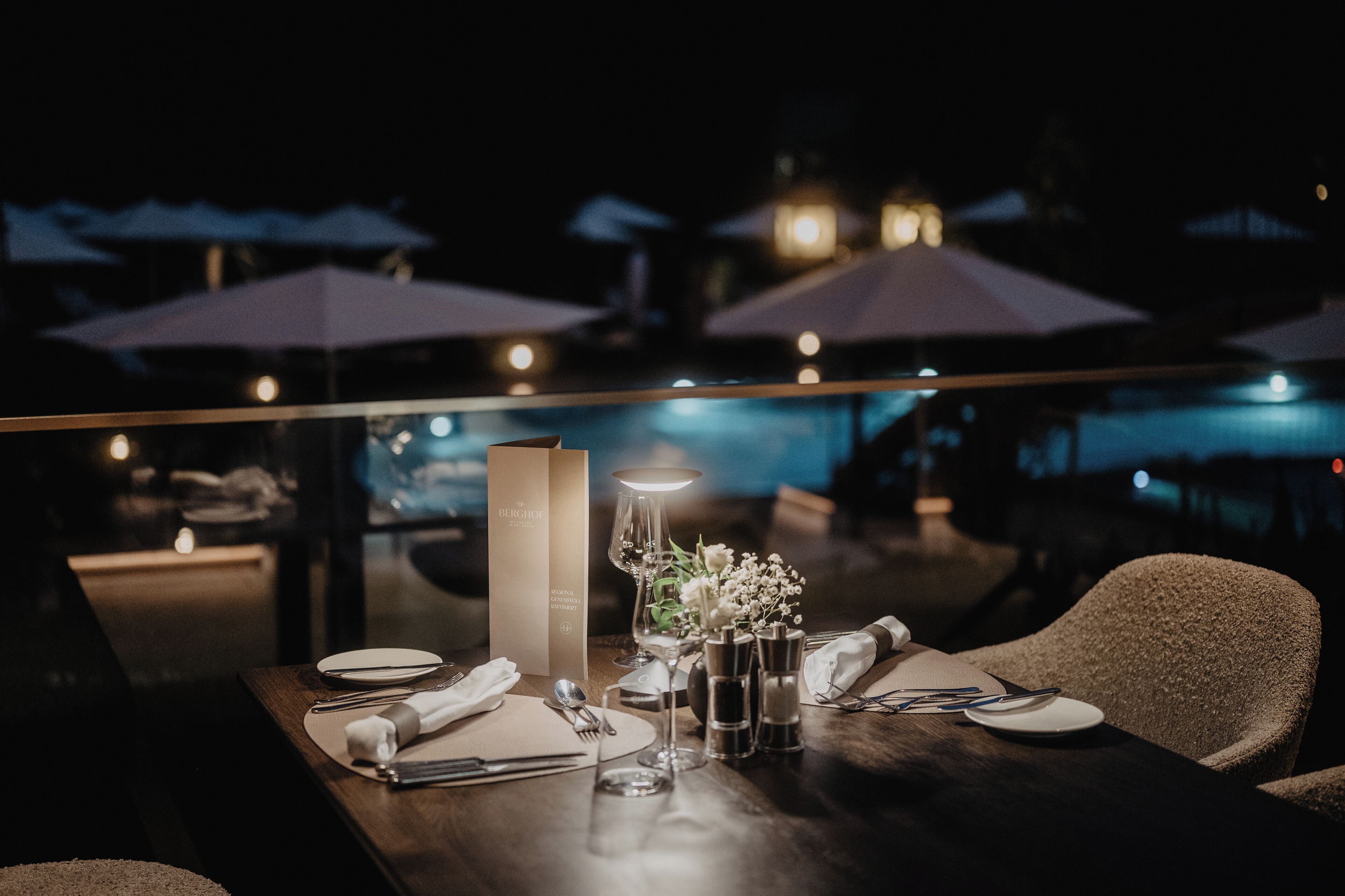 A dining table set for two with a menu, glasses, and a vase of flowers.