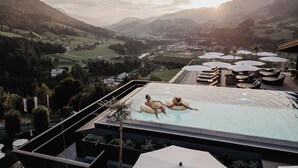 A rooftop pool with two people on inflatable rings, surrounded by lounge chairs and umbrellas, overlooking a valley with mountains in the background. -  Hotel Berghof | St. Johann in Salzburg (Sankt Johann im Pongau)