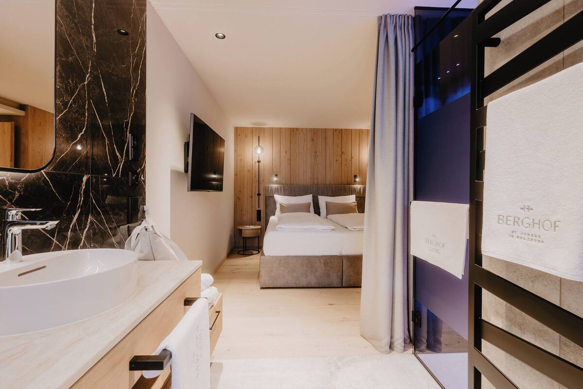 A modern hotel room with a large marble sink, a bed with a wooden headboard, a flat-screen TV, and a large mirror.