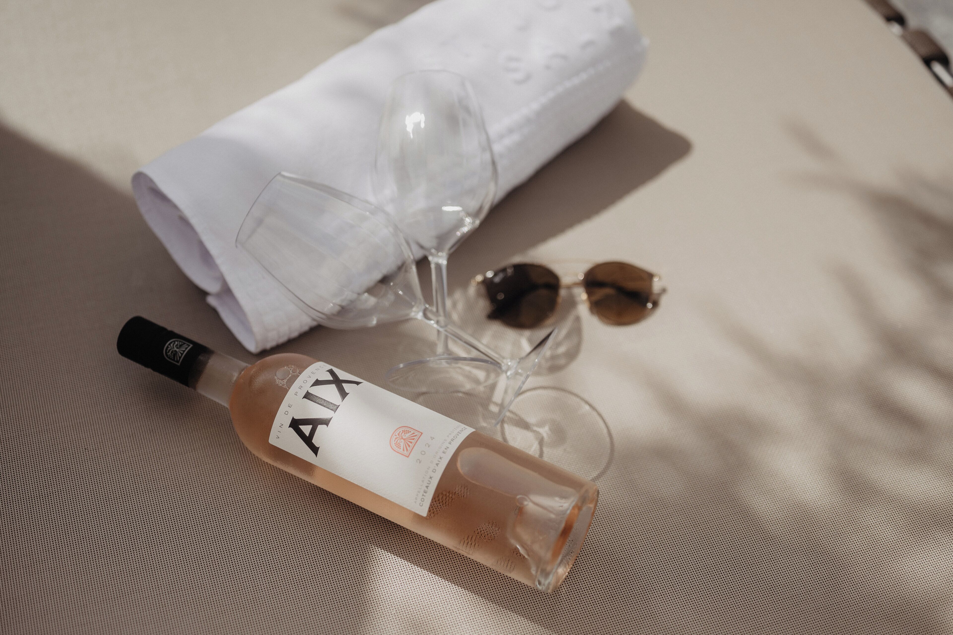 Une bouteille de vin AIX, une serviette blanche pliée et une paire de lunettes de soleil posées sur une surface texturée.