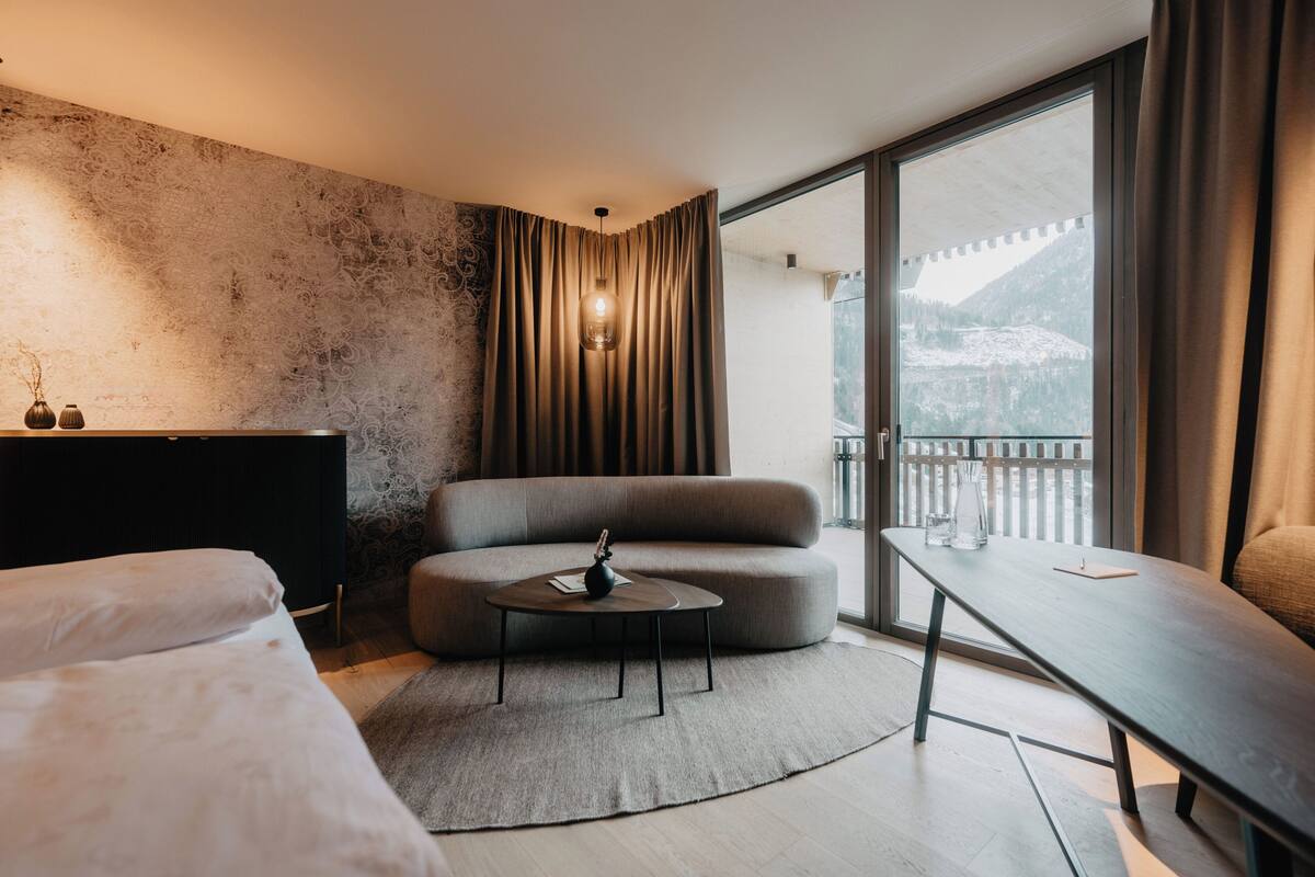 A modern hotel room with a large window, a sofa, a round table, and a bed.