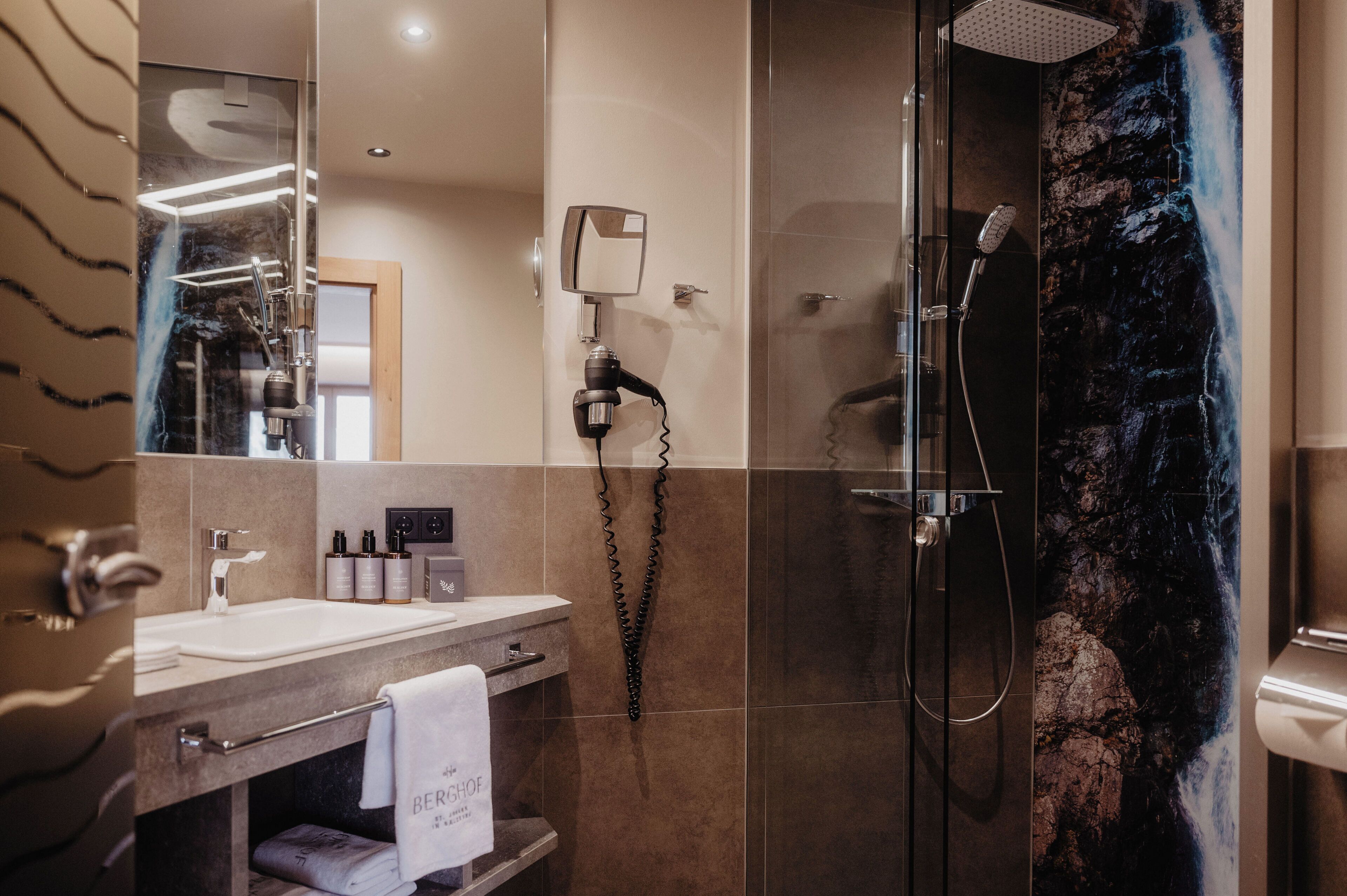 Et moderne badeværelse med et toiletbord, et stort spejl, en hårtørrer og en bruser med et vandfaldsmotiveret billedtryk.