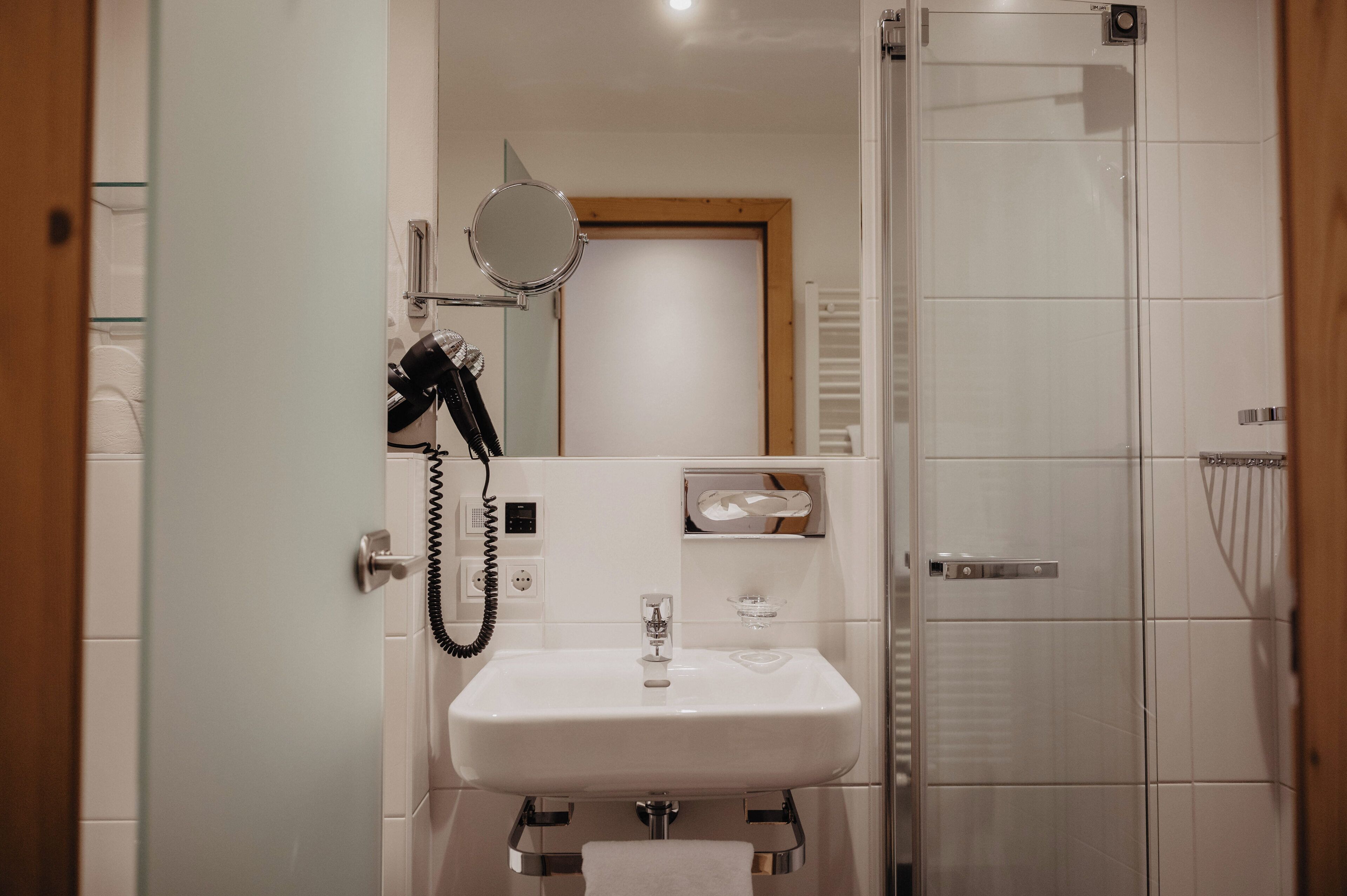 Ein kleines Badezimmer mit Waschbecken in Weiß, einem Spiegel, einer Duschbrause und einer Glasdusche.