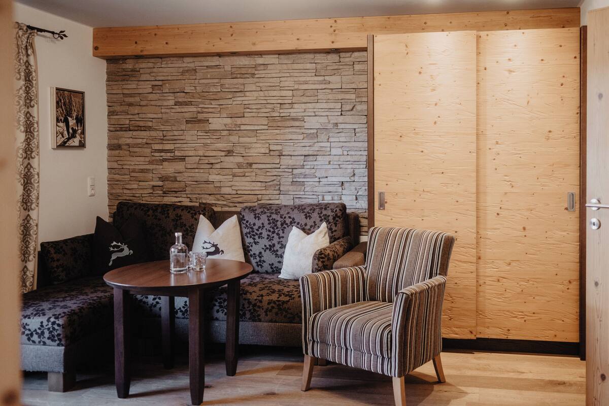 A cozy room with a wooden wall, a sofa, a striped armchair, and a small round table with a bottle and glasses.