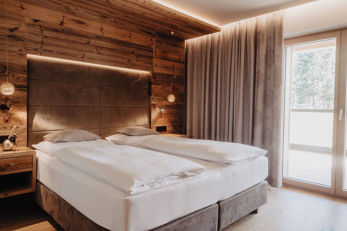 A modern bedroom with a wooden headboard, a large window with curtains, and a bedside table with a lamp.