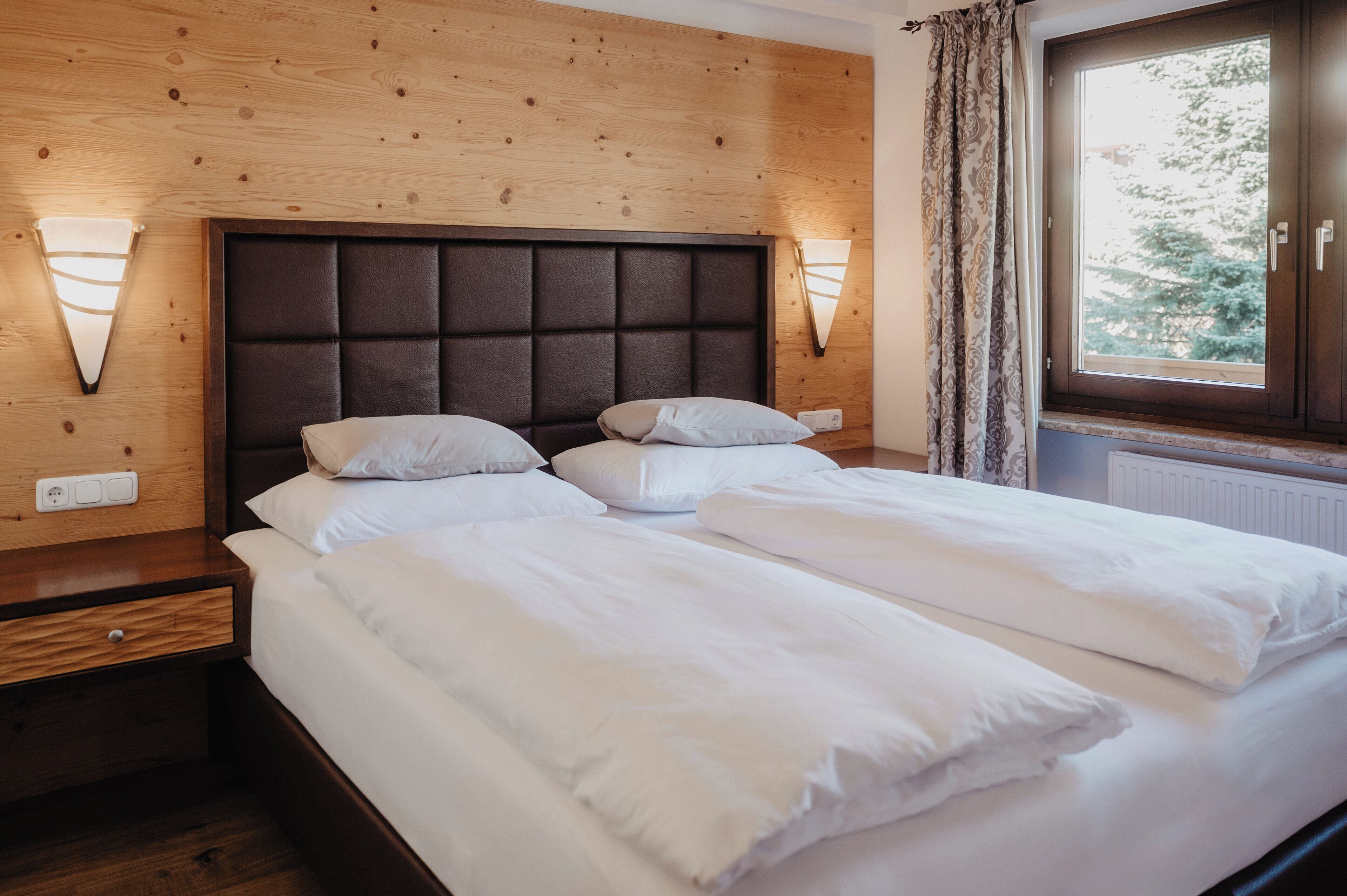 A neatly made bed with white linens in a room with wooden paneling and a large window with curtains.