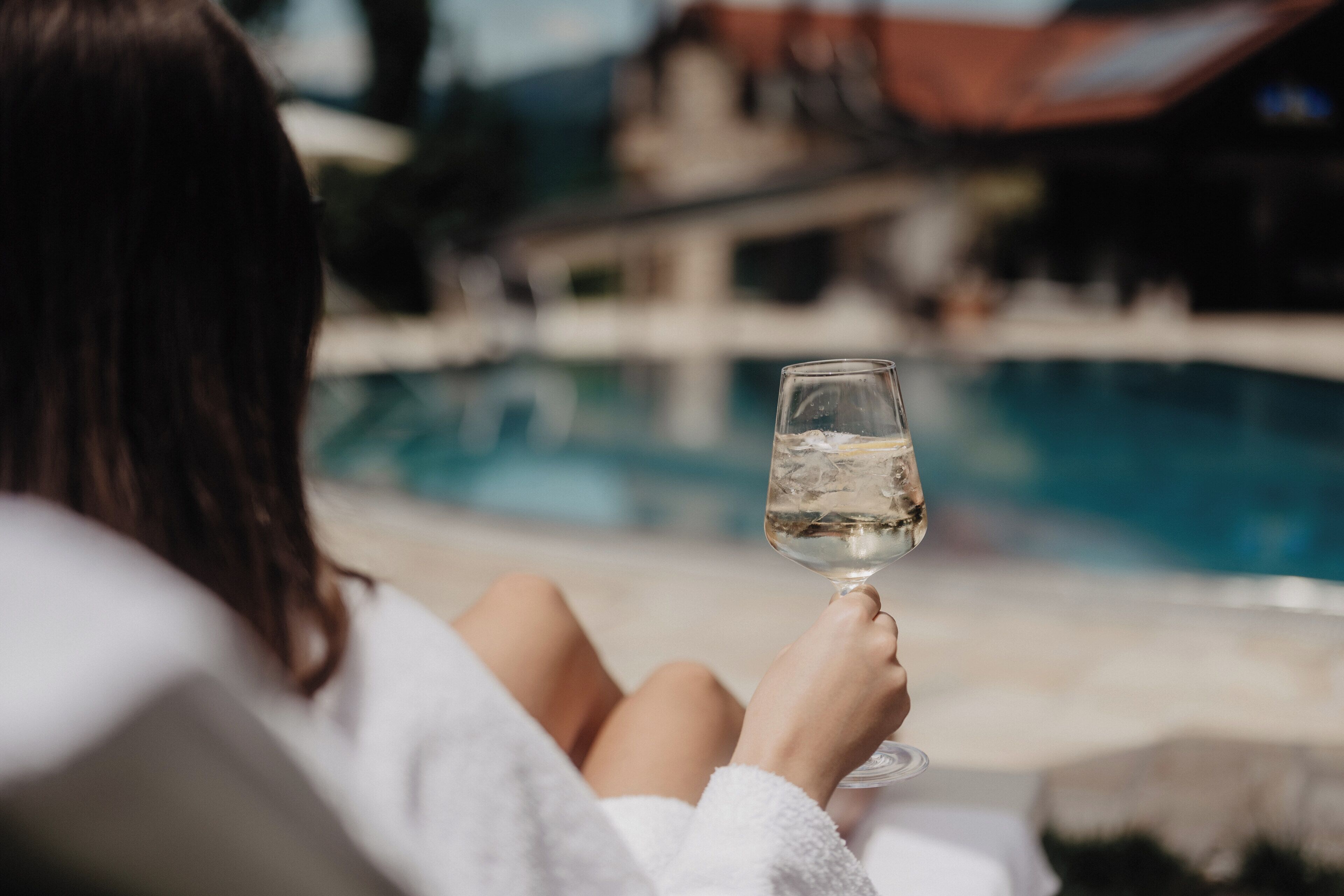A person relaxing by a pool with a glass of water, wearing a white robe.