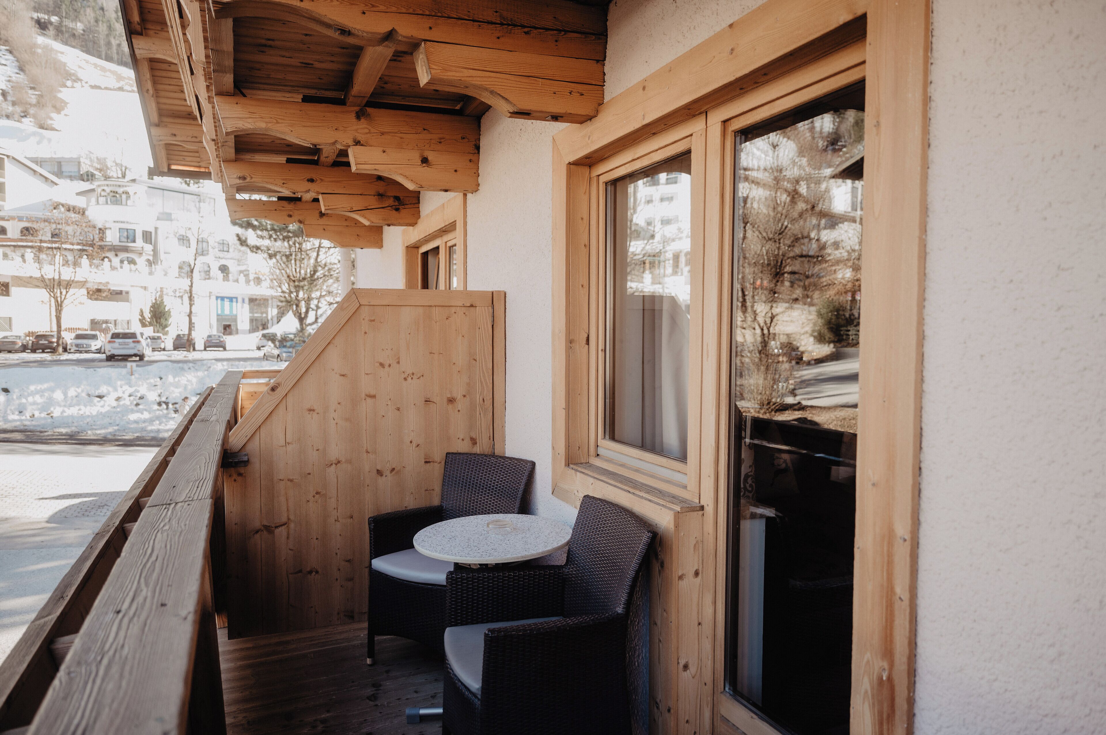 Un balcón de madera con sillas de mimbre y una pequeña mesa redonda, con vistas a una calle nevada.