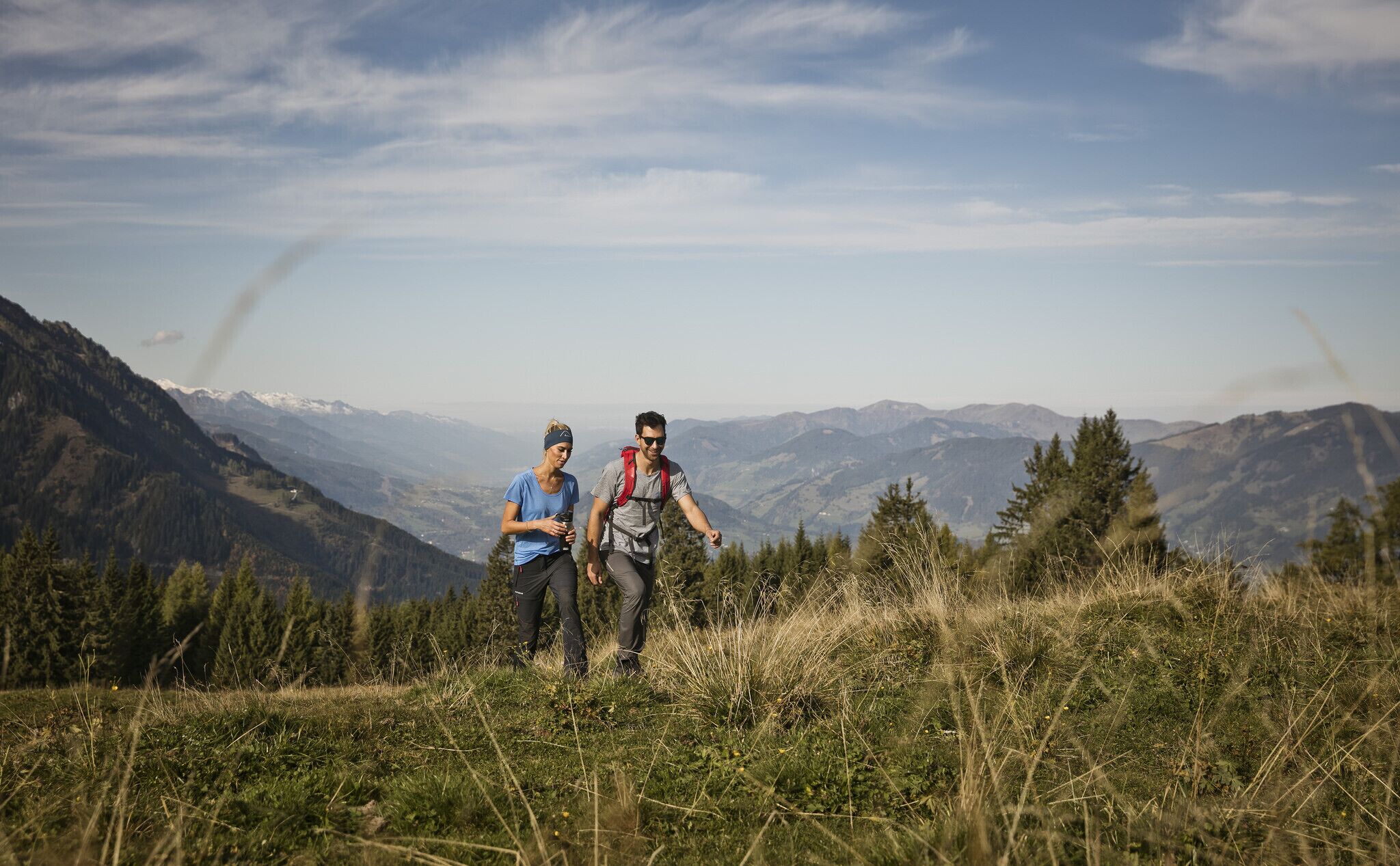 Two people hiking on a grassy mountain trail with a scenic mountain view in the background.