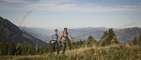 Two people hiking on a grassy mountain trail with a scenic mountain view in the background.