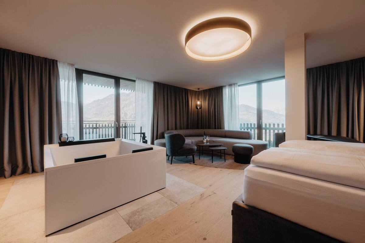 A modern hotel room with a large bed, a sitting area with a sofa and chairs, a small table, a bathtub, and large windows with curtains.