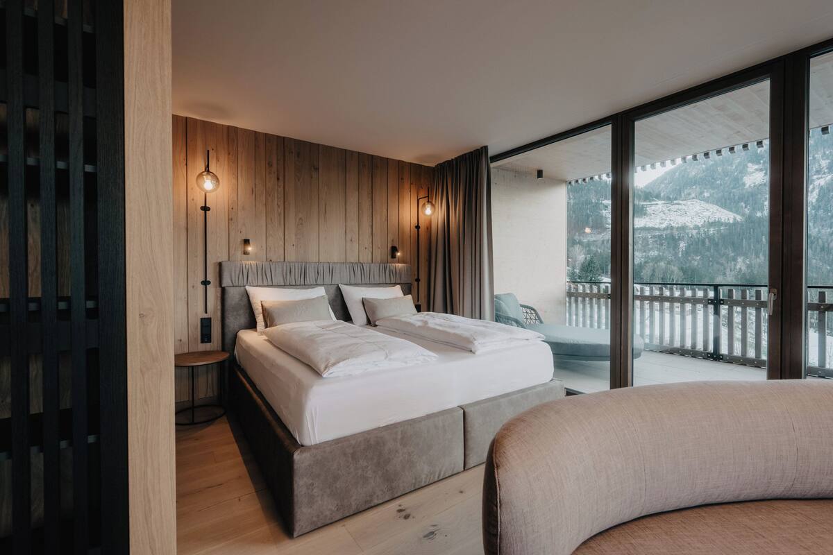 A modern bedroom with a large bed, wooden panel walls, and a balcony with a mountain view.