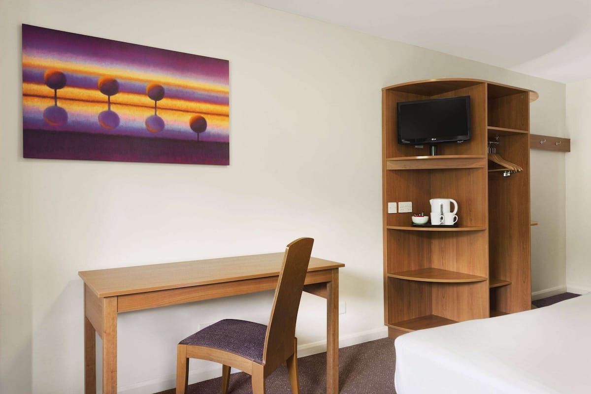 Une chambre d’hôtel avec un bureau en bois, une chaise, un lit, un téléviseur posé sur un meuble en bois et un tableau accroché au mur.