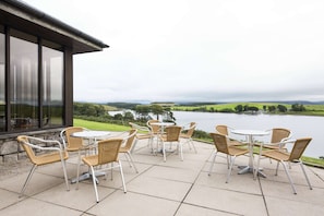 Pátio externo com cadeiras de vime e uma mesa, com vista para um lago e colinas ao longe.