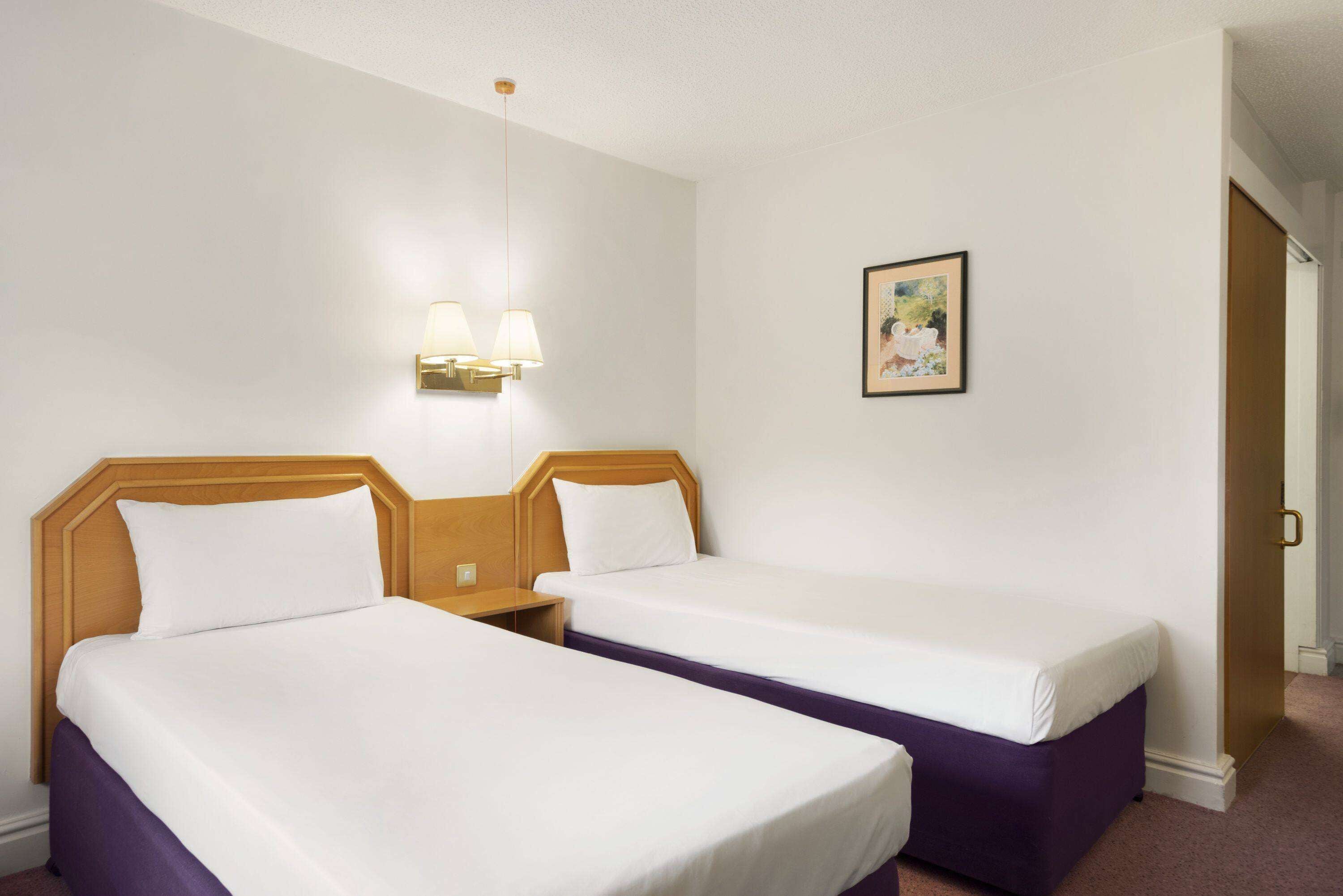 Habitación de hotel con dos camas individuales, una mesita de noche, una lámpara de pared y un cuadro enmarcado.