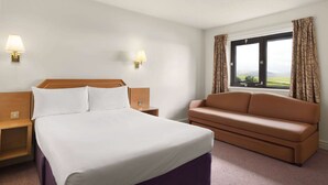 Une chambre d’hôtel comprenant un lit, une table de chevet, un canapé, deux lampes fixées au mur et une fenêtre avec des rideaux.