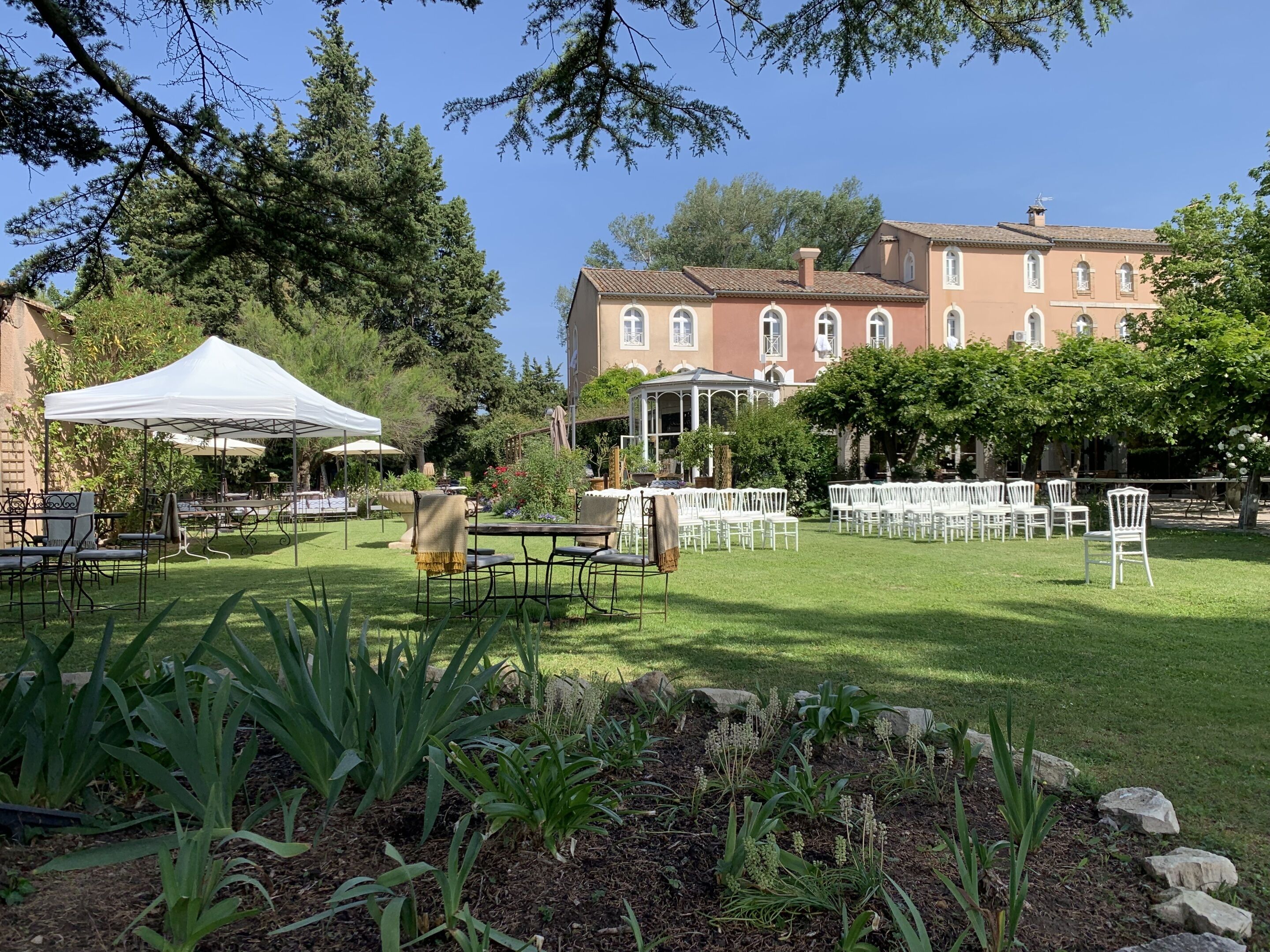 An outdoor garden with tables and chairs, a white tent, and a building in the background.