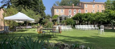 An outdoor garden with tables and chairs, a white tent, and a building in the background.