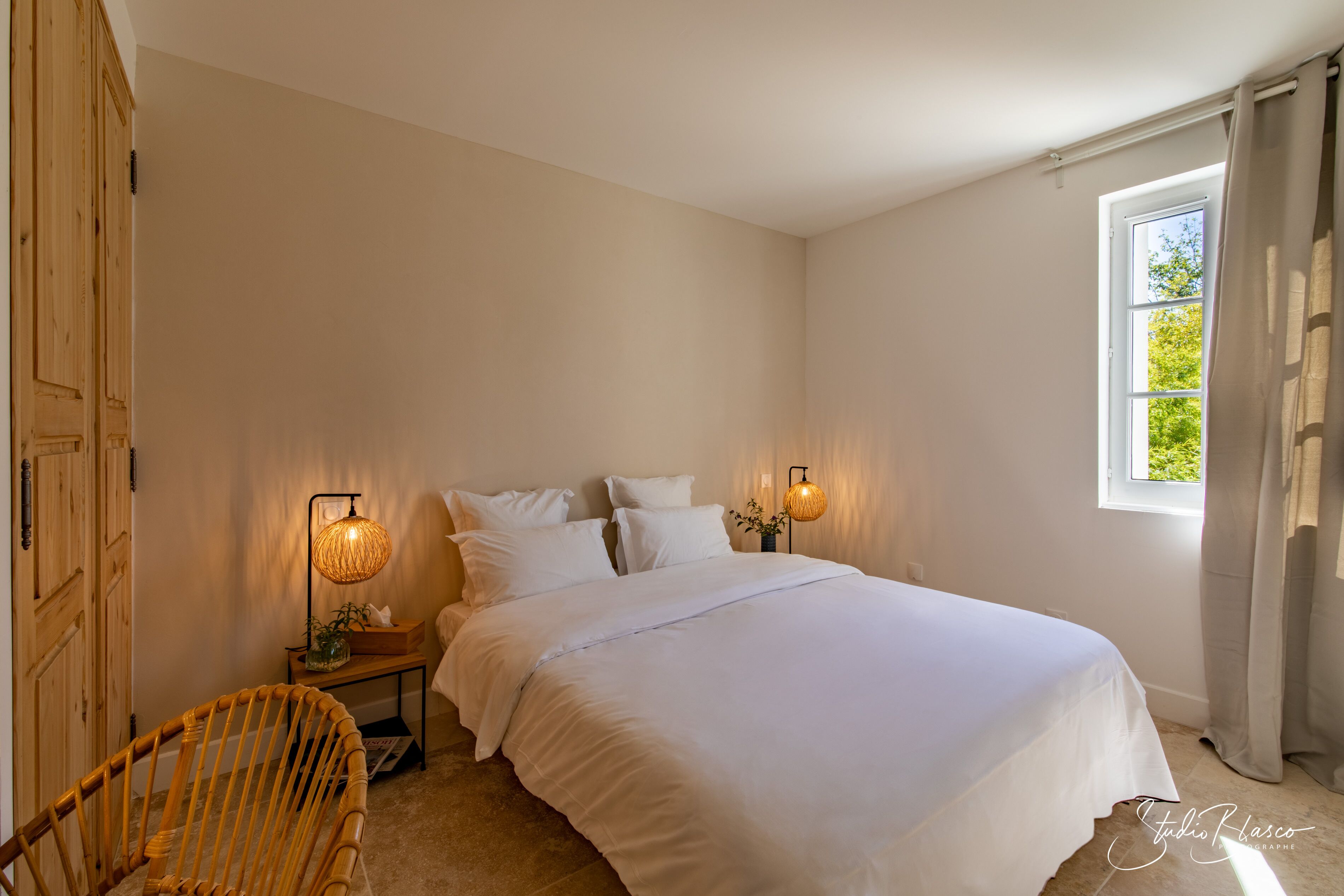A bedroom with a bed, two bedside lamps, a wooden wardrobe, a chair, and a window with curtains.