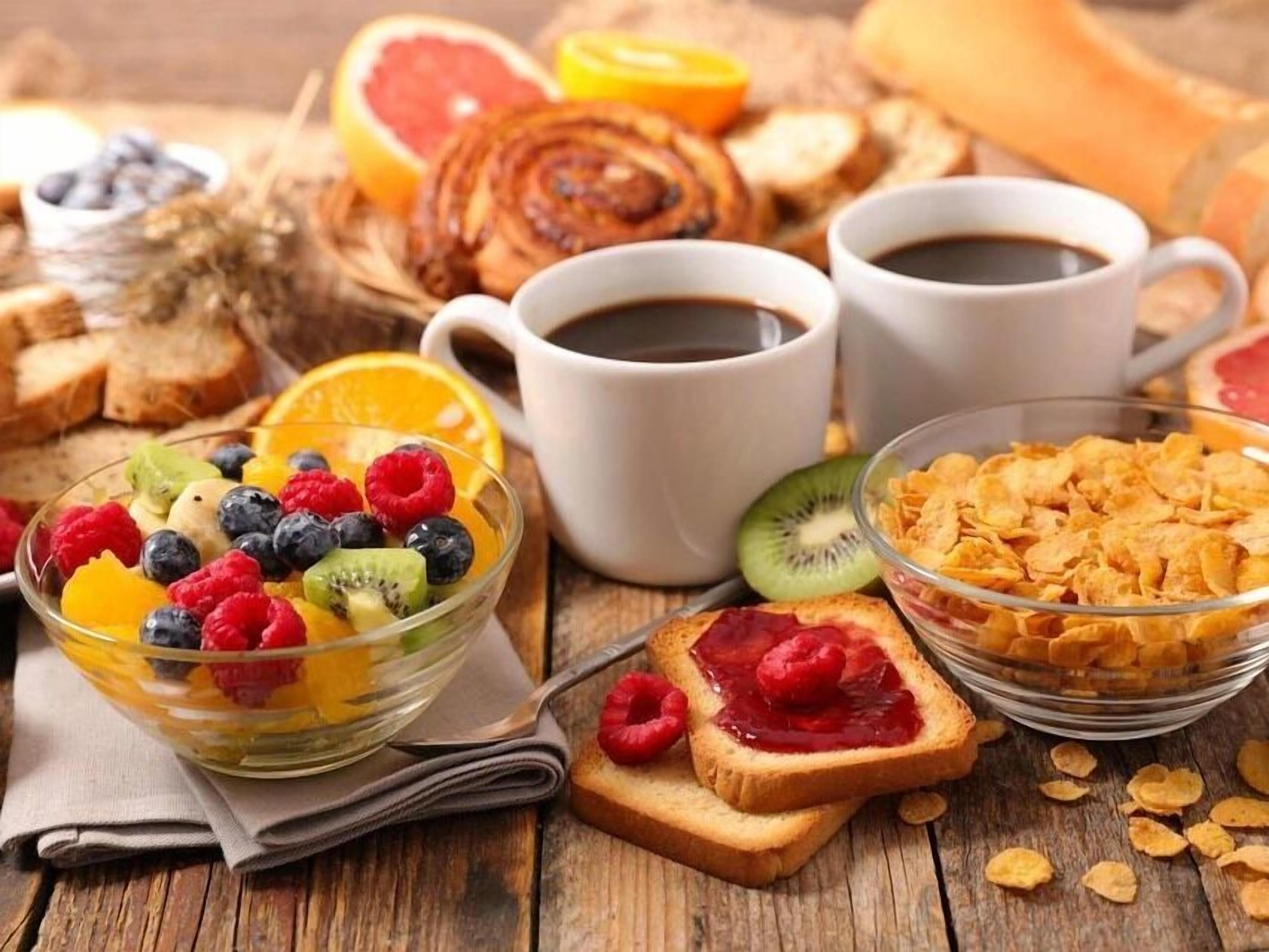 Une table en bois dressée pour le petit-déjeuner, avec un bol de fruits variés, des tartines à la confiture, des céréales et deux tasses de café.