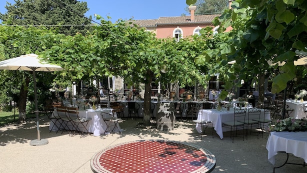 Un área de comedor al aire libre con mesas y sillas redondas, rodeada de follaje y un edificio en el fondo.