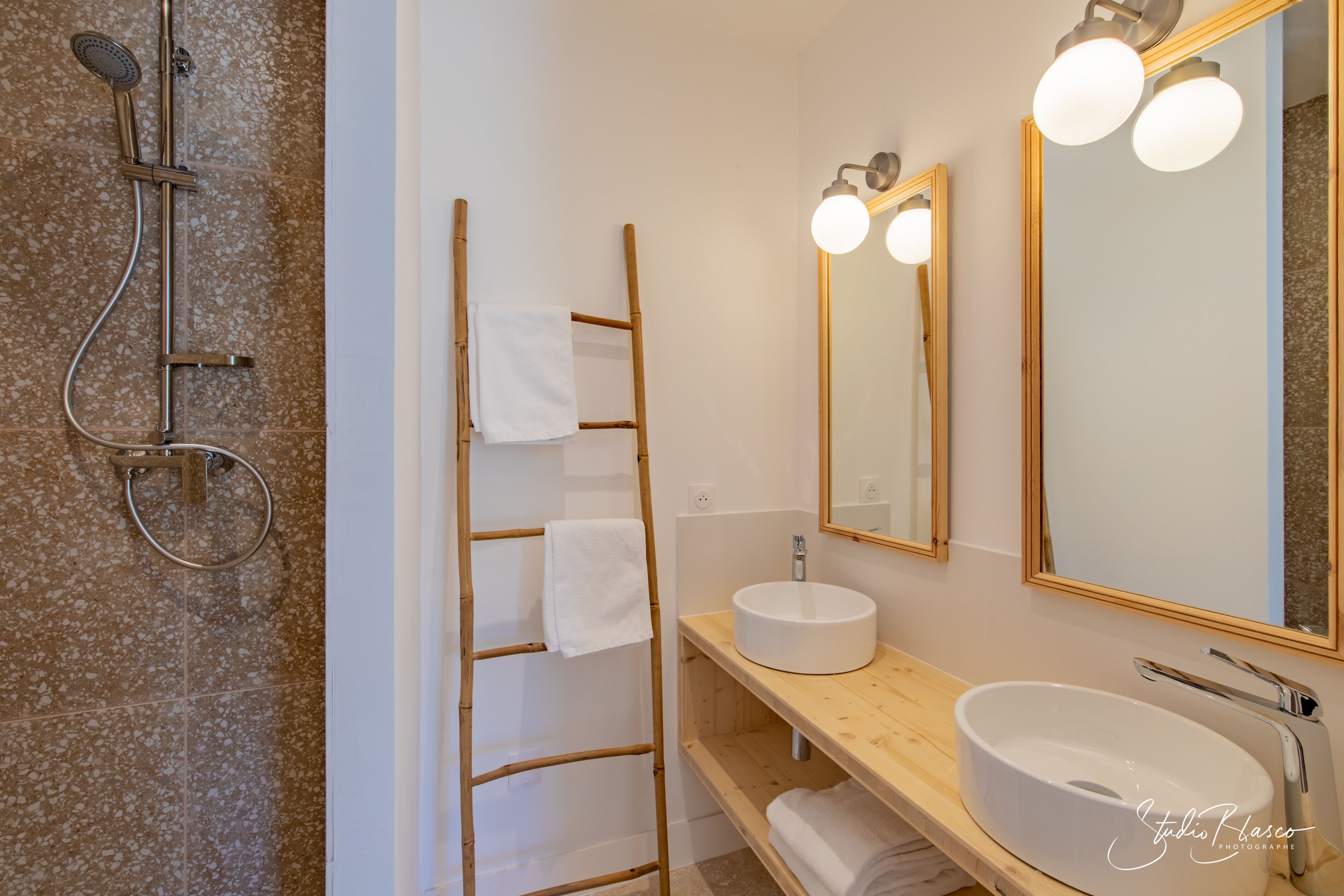 A bathroom with a freestanding shower, a wooden ladder, a round sink, and a large mirror with lights.