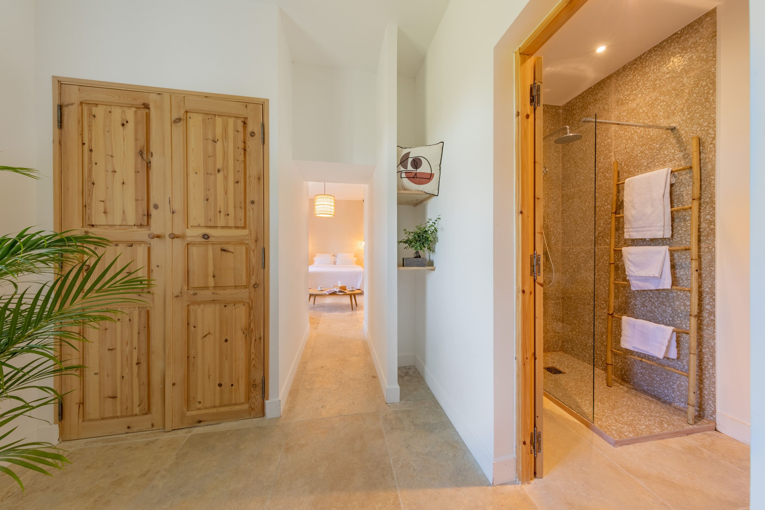 A hallway with wooden doors, a glass shower area, and a bed in the background.