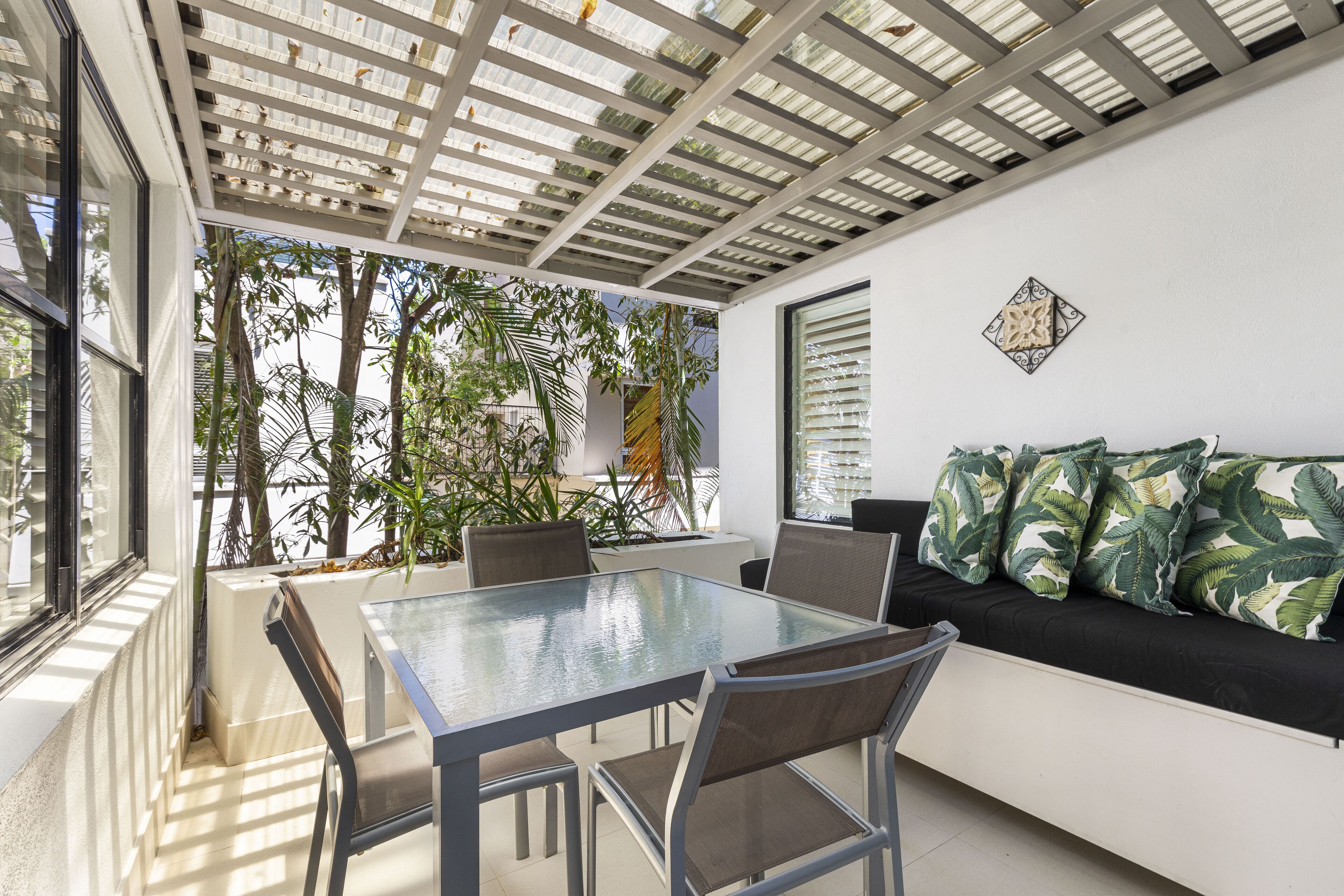 Ein zeitgemäßer Essbereich im Freien mit einem Glastisch und Stühlen, einer schwarzen Couch mit tropischen Kissen und einer Pergola darüber.