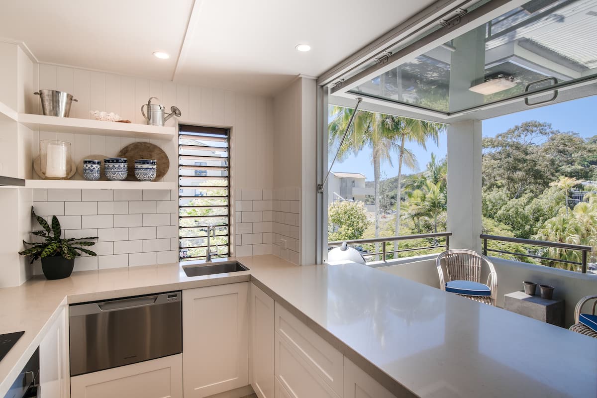 A modern kitchen with white cabinetry, a built-in dishwasher, and a large window overlooking a balcony with a view of palm trees and a hillside.