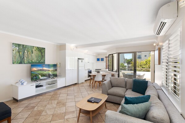 A modern living room with a flat-screen TV, a sofa, a coffee table, and a dining area with chairs.
