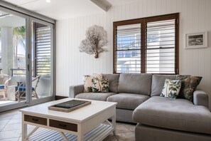 A living room with a sofa, a coffee table, and a sliding glass door leading to an outdoor area with chairs. - Bella Casa Noosa (Noosa Heads)