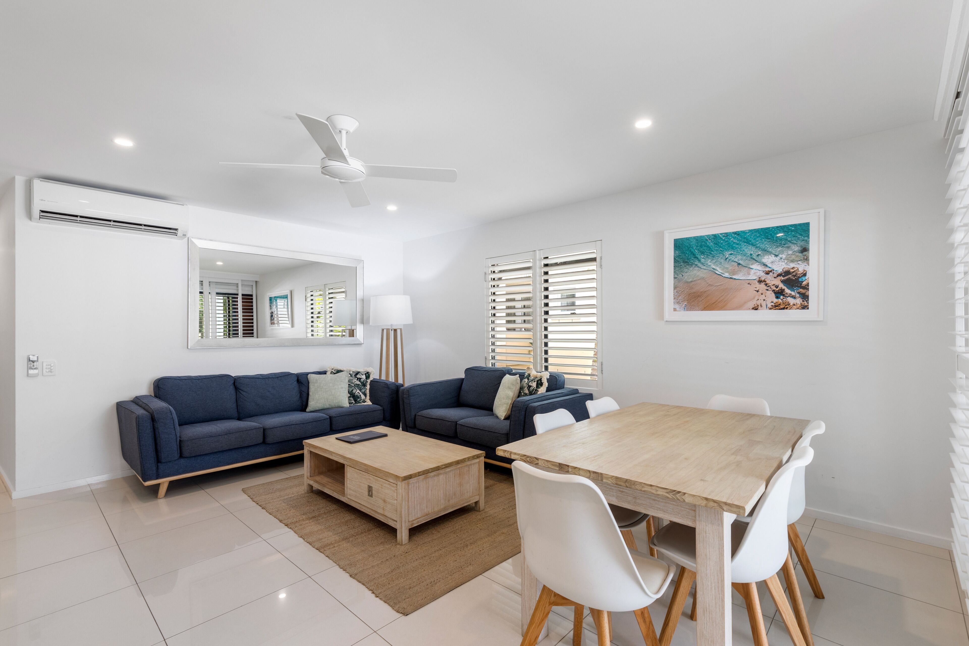 A modern living room with a blue sofa set, a wooden dining table with white chairs, a ceiling fan, and a beach-themed painting on the wall.