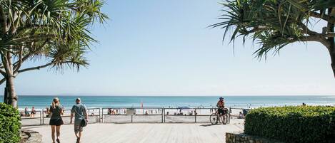 A paved walkway with palm trees, a beach, and a cyclist.