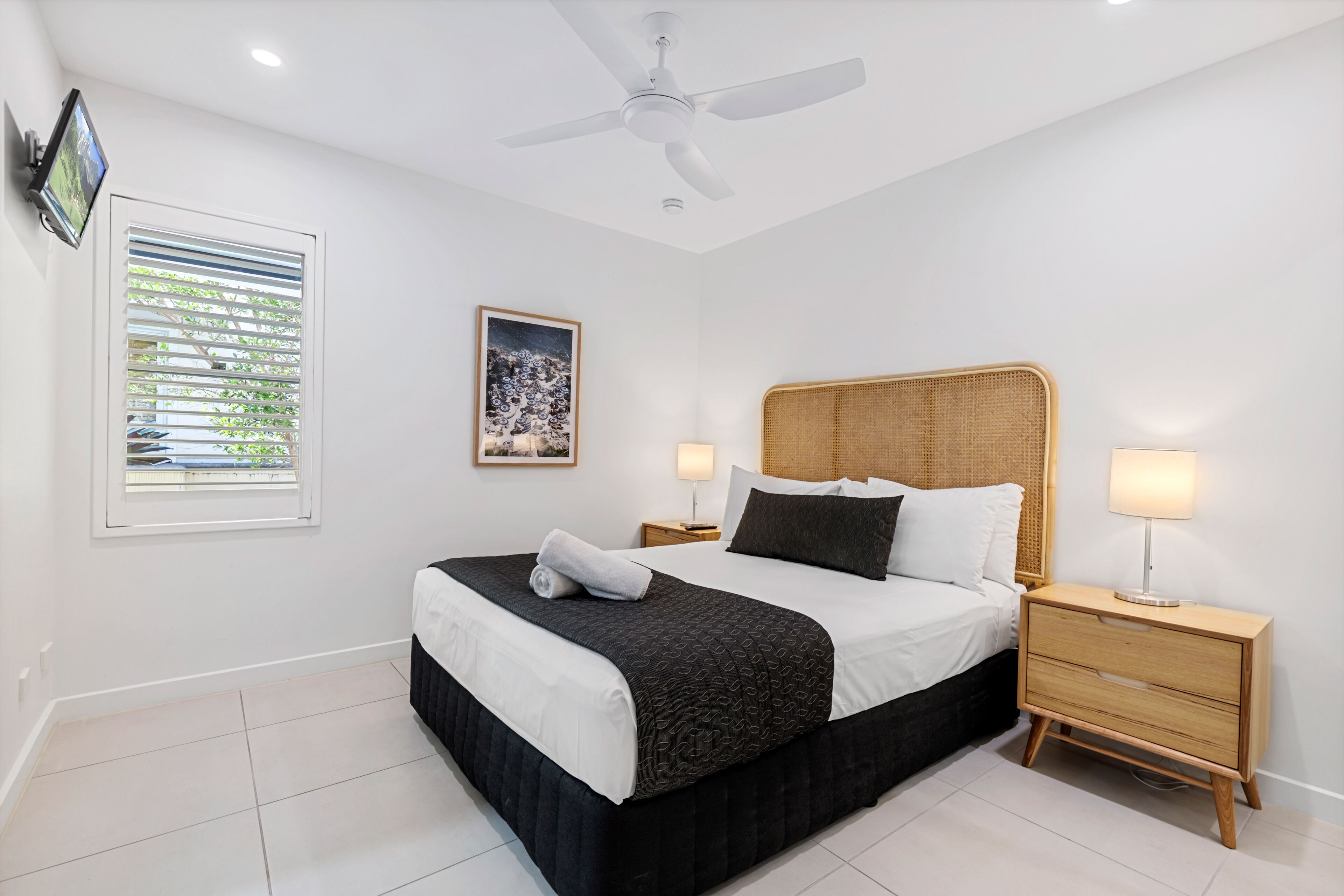 A bedroom with a bed, bedside lamps, a ceiling fan, a window with blinds, and a framed picture on the wall.