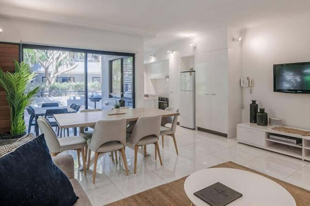 A modern kitchen with white cabinets, a dining area with a table and chairs, and a flat-screen TV mounted on the wall.