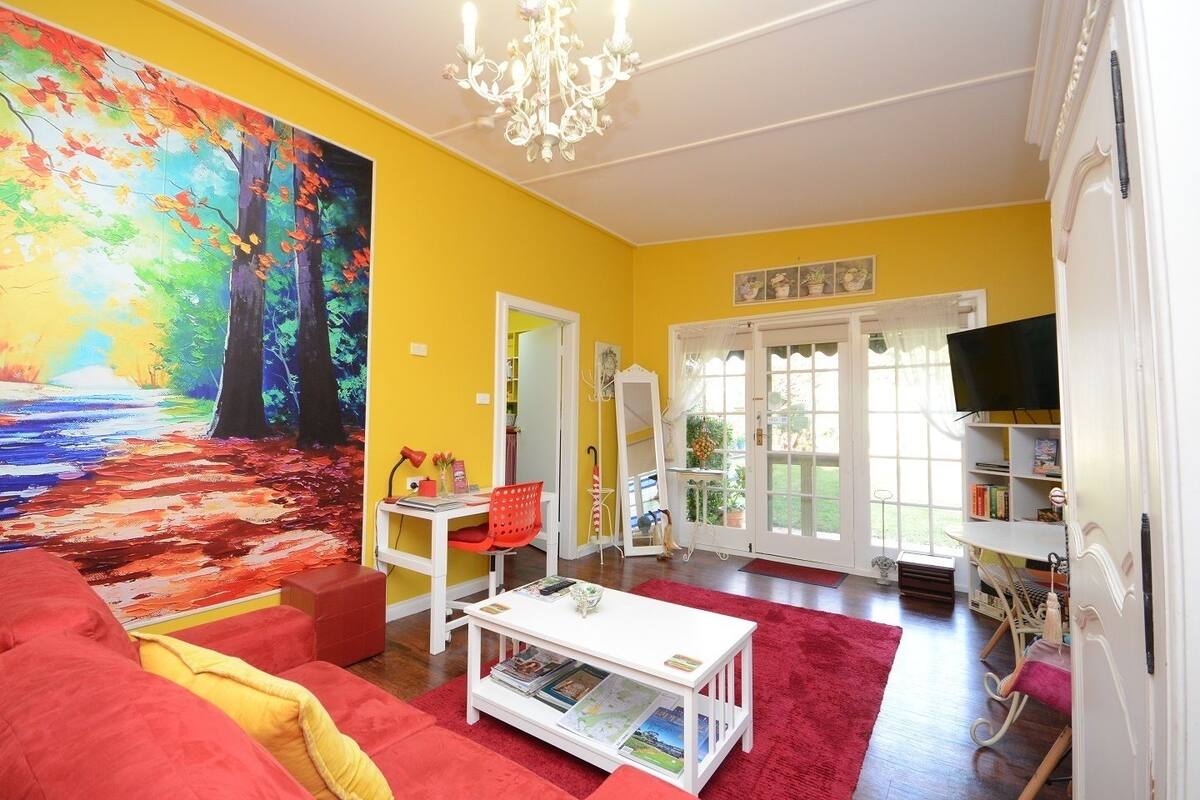 Een woonkamer met een levendige gele wand, een groot schilderij van een bos, een rode bank, een witte bijzettafel en een televisie op een witte standaard.