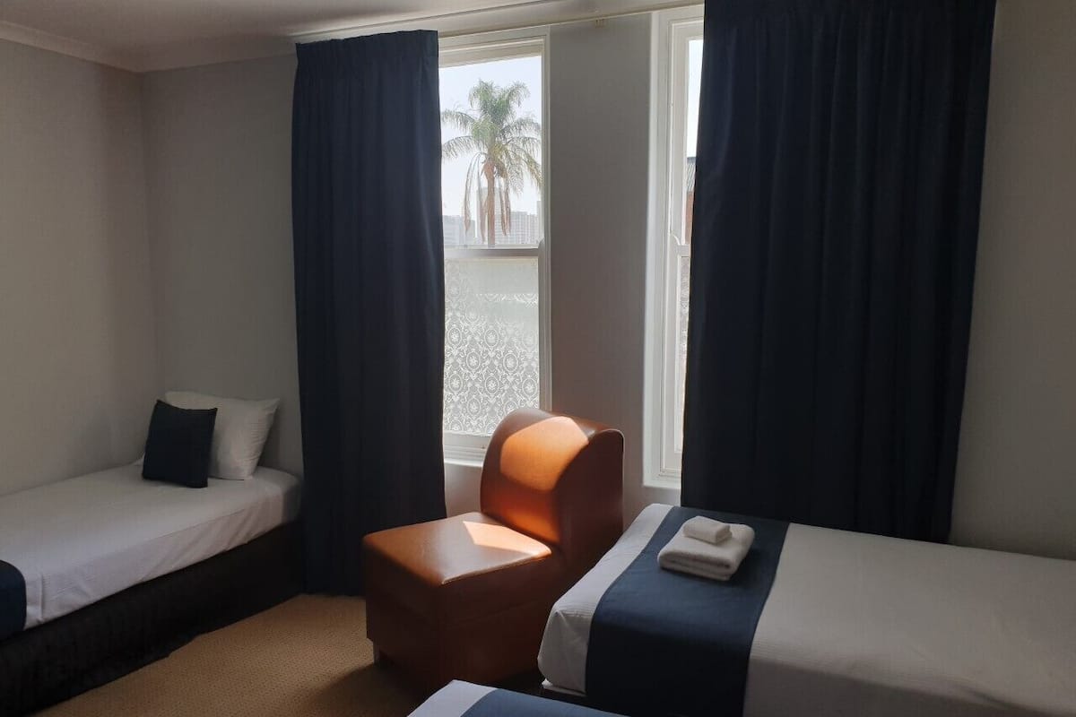 A hotel room with two single beds, a small armchair, and a window with curtains.