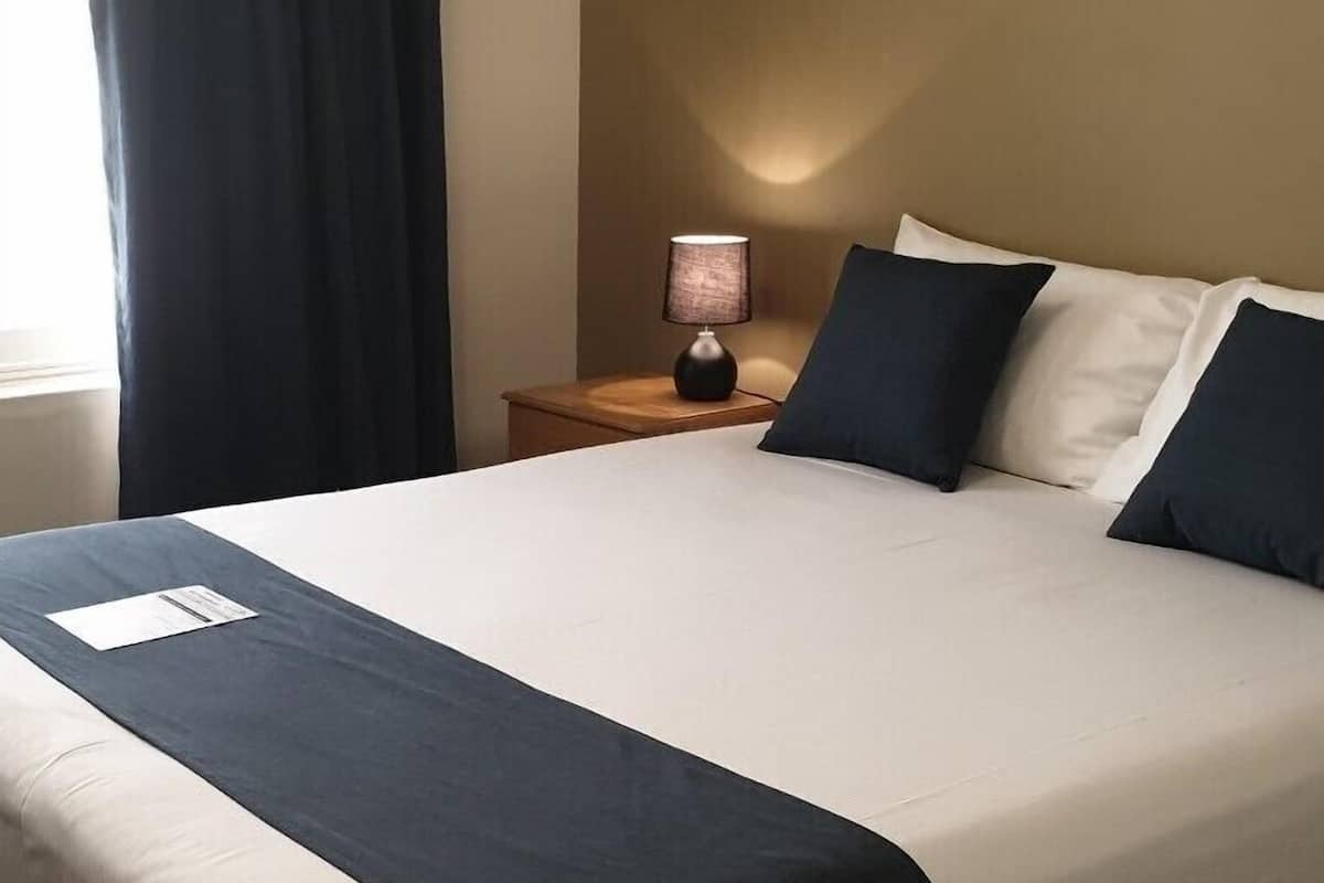 A neatly made bed with a white and dark blue bedspread, a small bedside table with a lamp, and a dark curtain partially drawn.