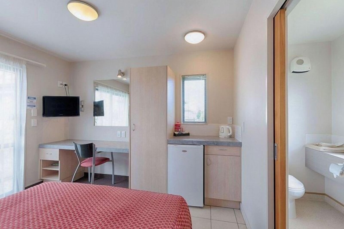 A compact hotel room with a bed, a desk with a chair, a small kitchenette, a television, and a bathroom.