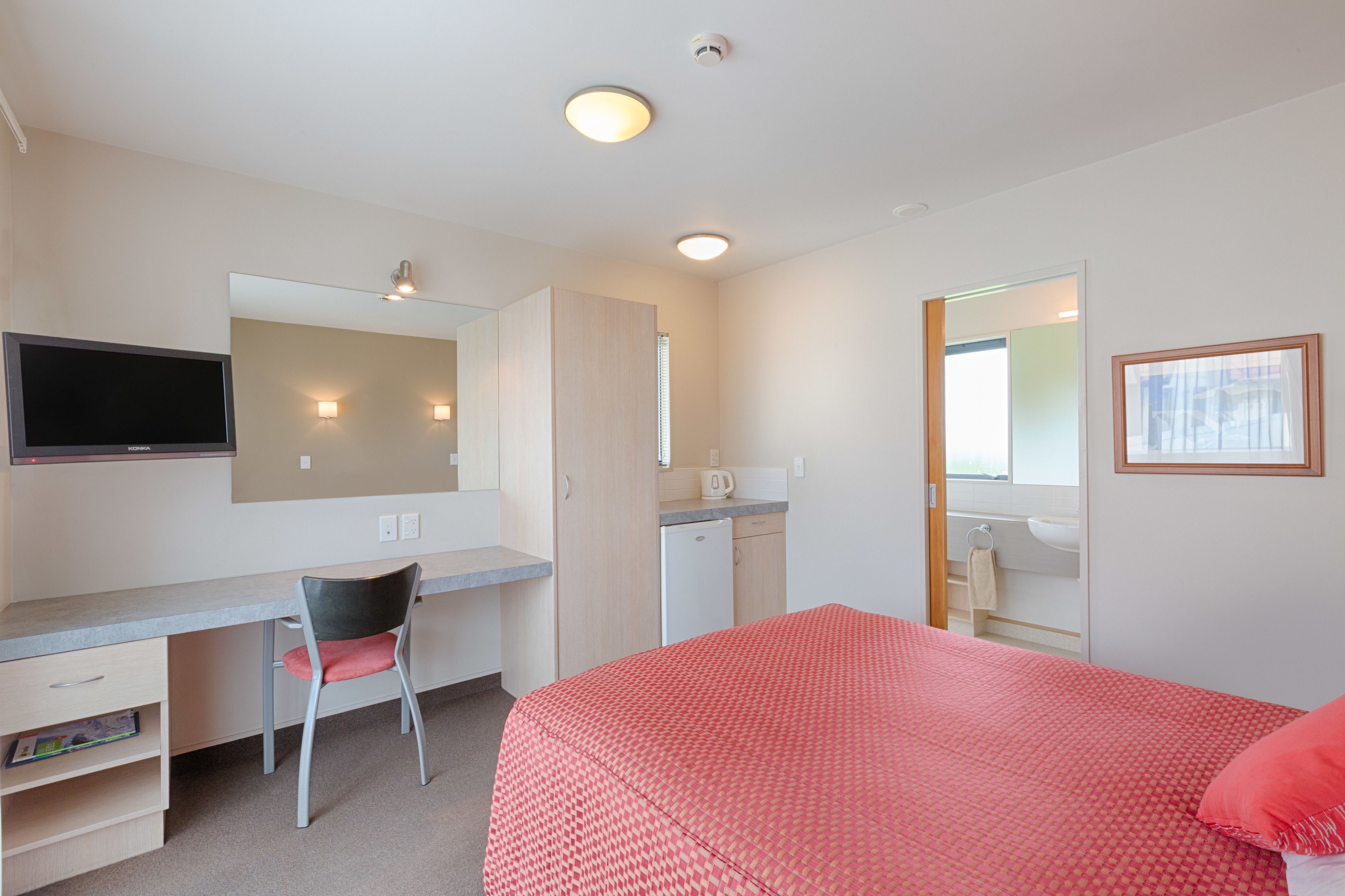 A hotel room with a bed, a desk with a chair, a television, a mirror, and a bathroom.