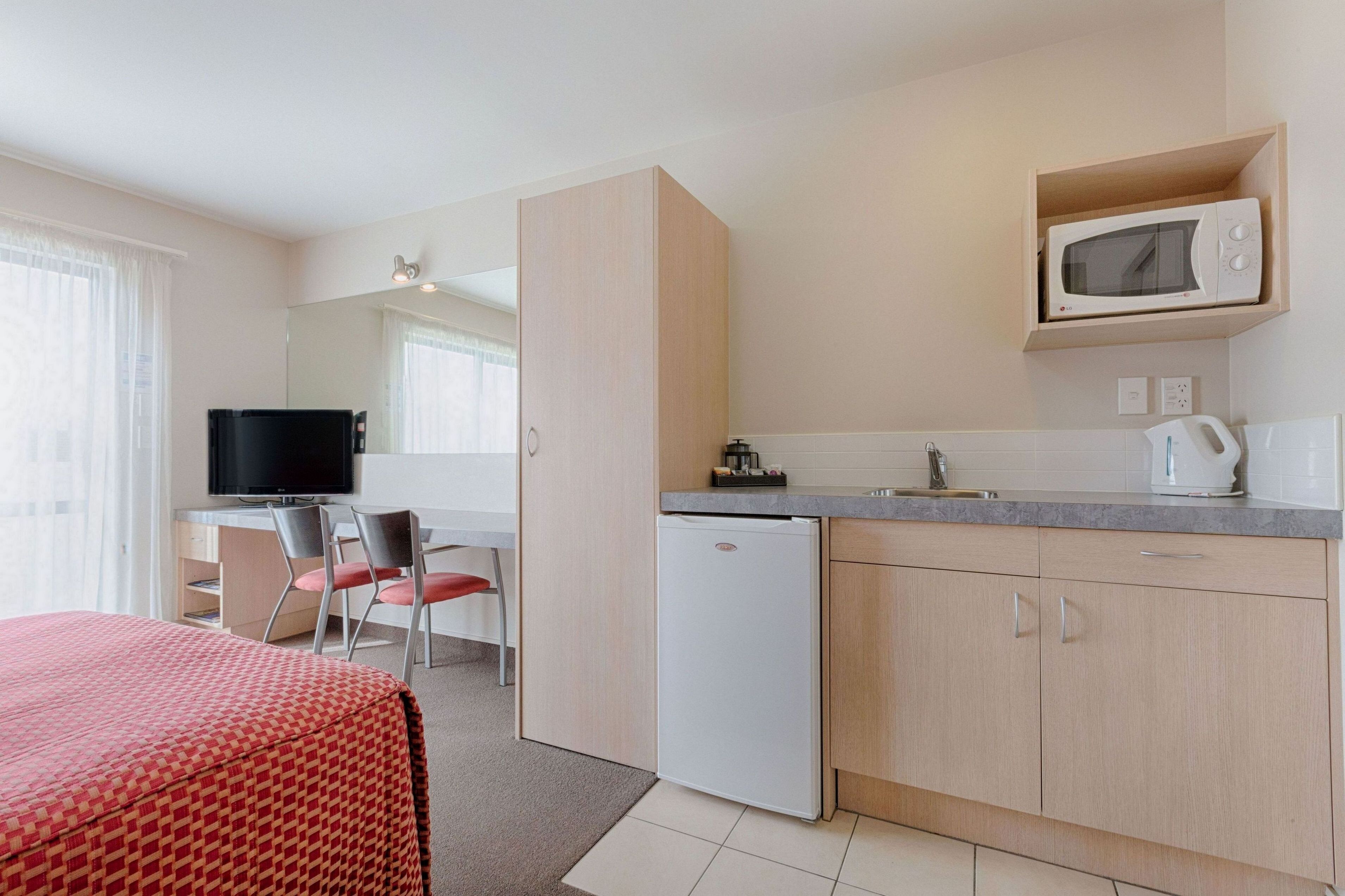 A compact hotel room with a kitchenette, a bed with a red patterned cover, a television, and a small refrigerator.