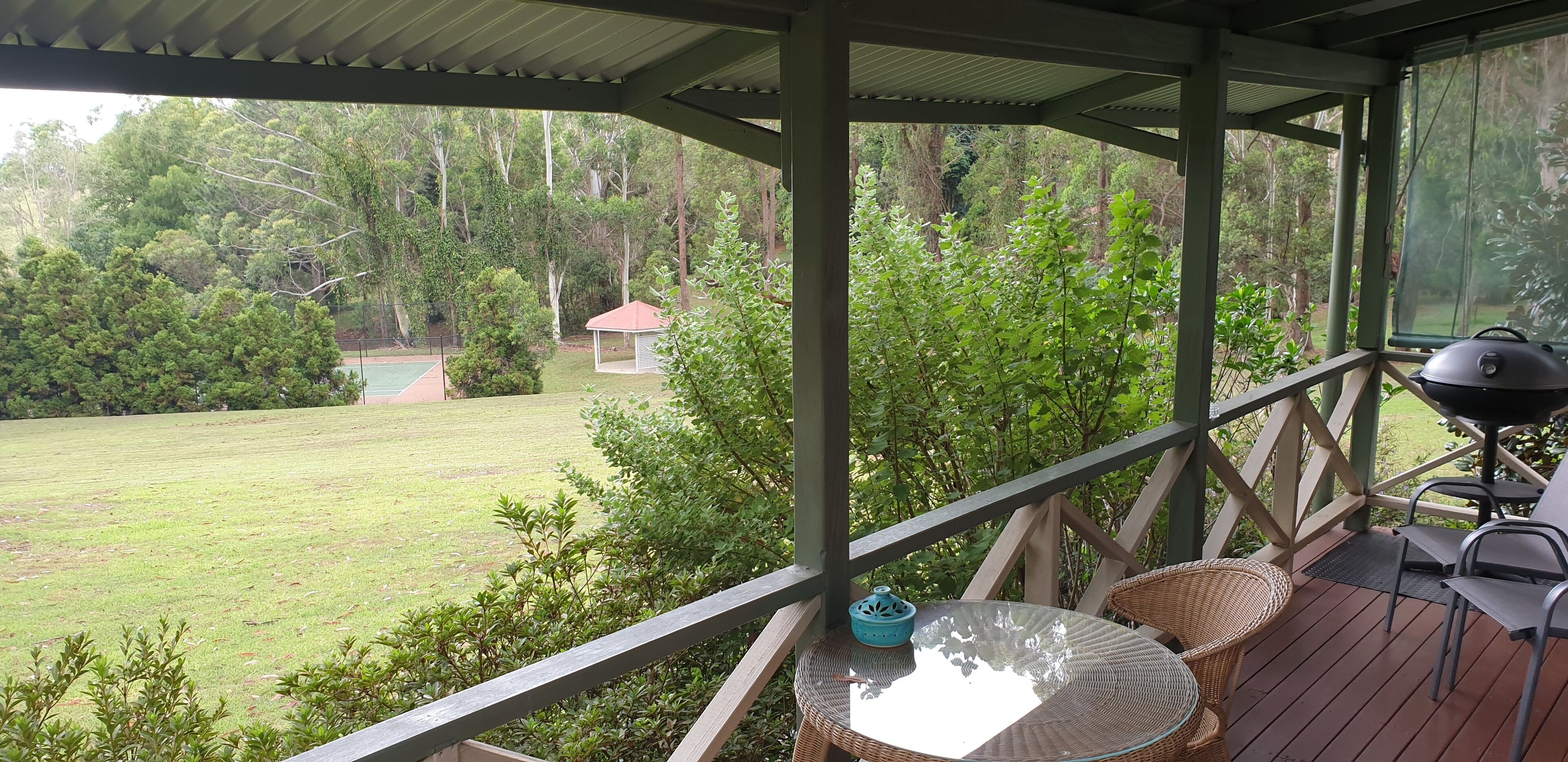 Un espace extérieur abrité, équipé d’une table et de chaises, d’un barbecue et offrant une vue sur un champ verdoyant et des arbres.